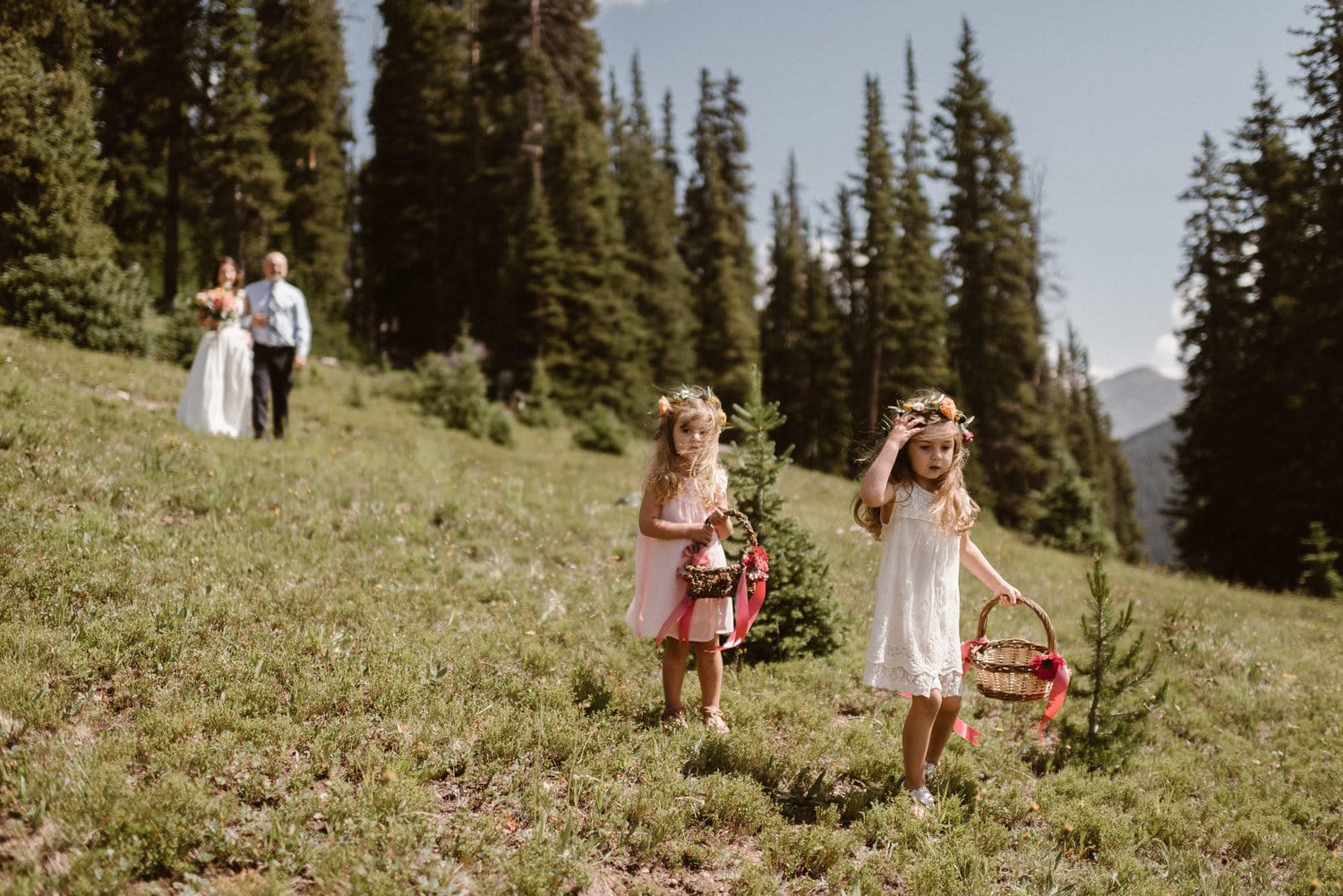 Two young girls wearing flower crowns and holding baskets, walking in front of bride and her father. There are pine trees in the background.