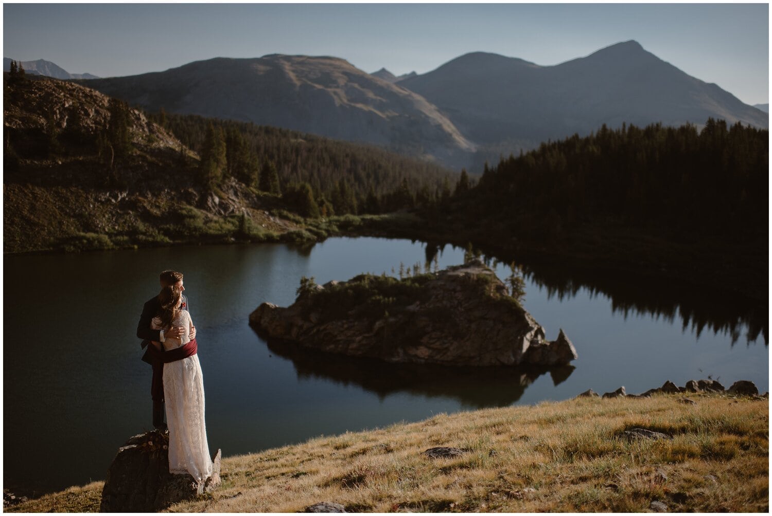 Bride and groom hugging while standing on rock in front of alpine lake, with mountains and trees in background. 