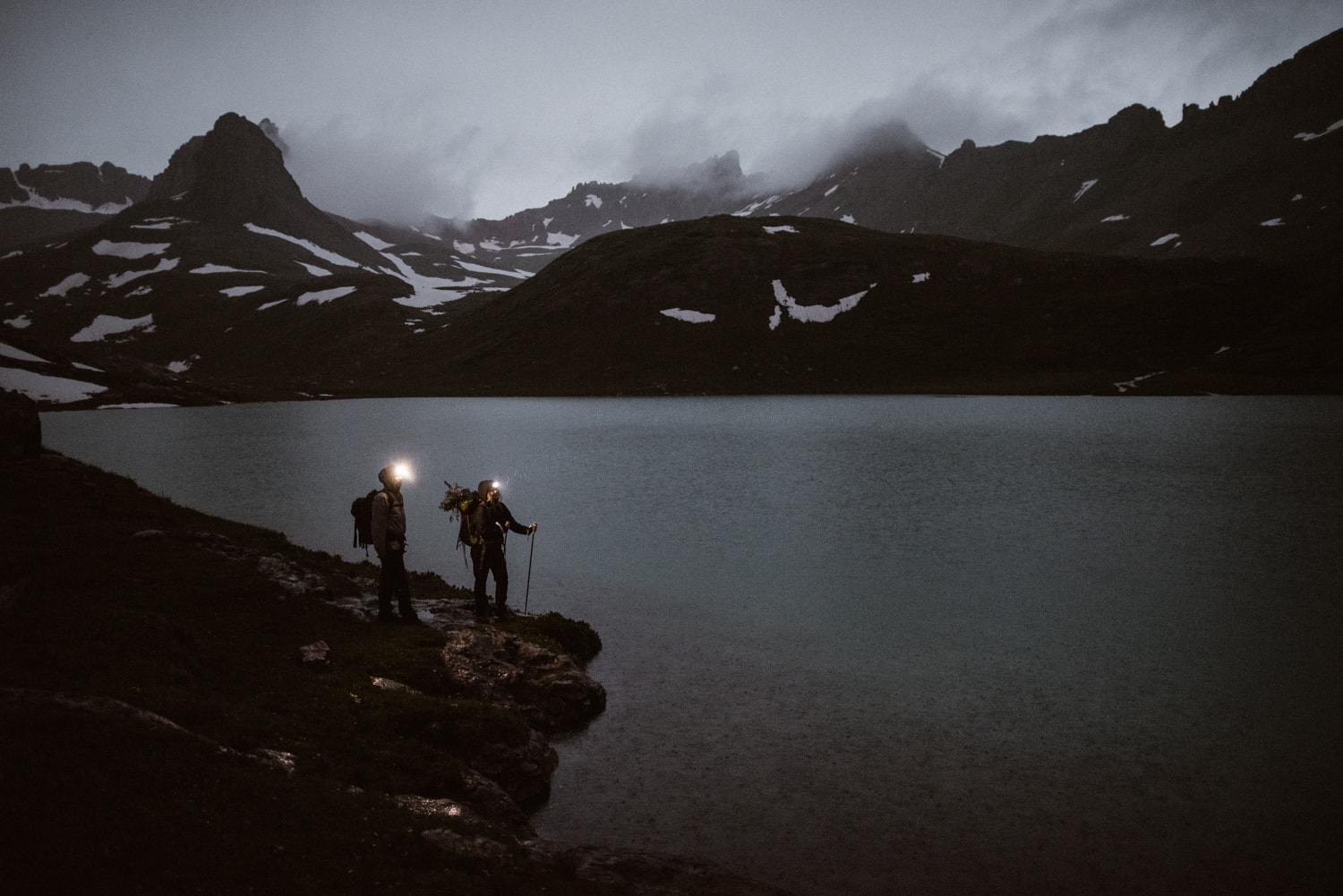 Bride and groom standing at the edge of an alpine lake in the dark. They are wearing headlamps and there are snow-covered mountains in the background. 