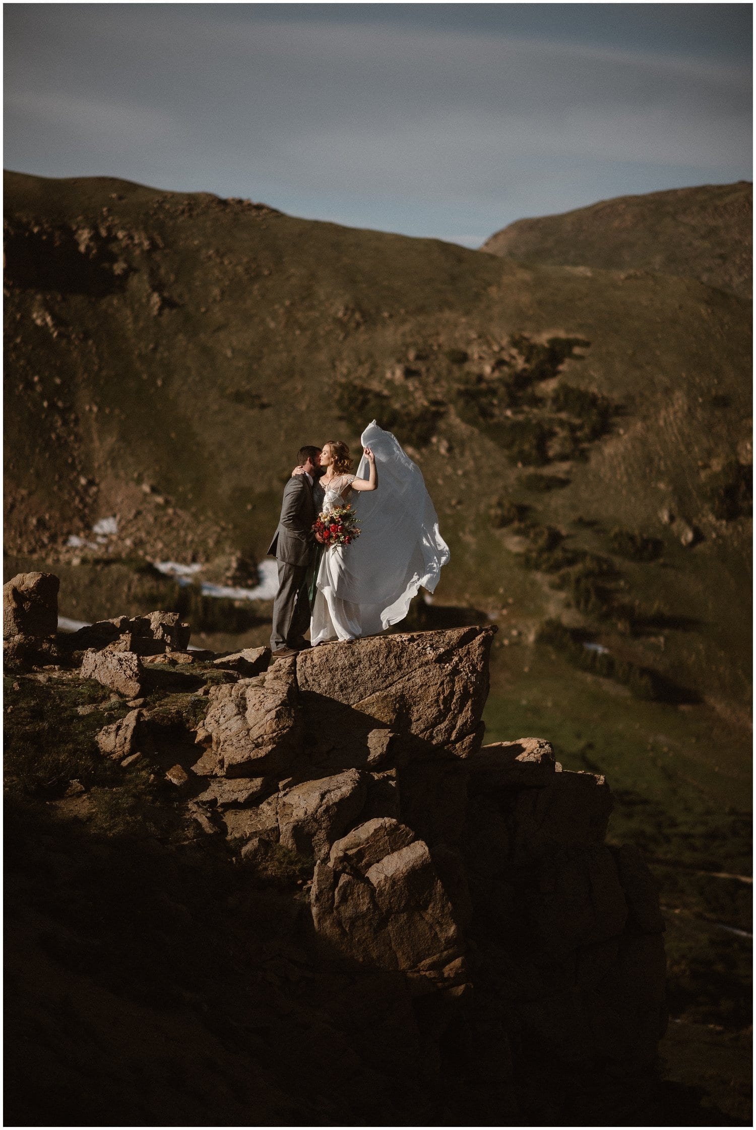 Bride and groom embrace on a cliff on their elopement day. The bride's dress flows in the wind behind her. 