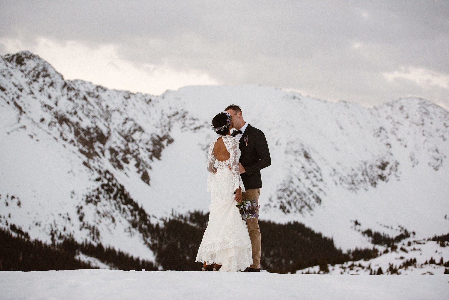 Bride and groom kiss at Loveland Pass, in Colorado. There is snow covering the ground and the mountains in the background.  
