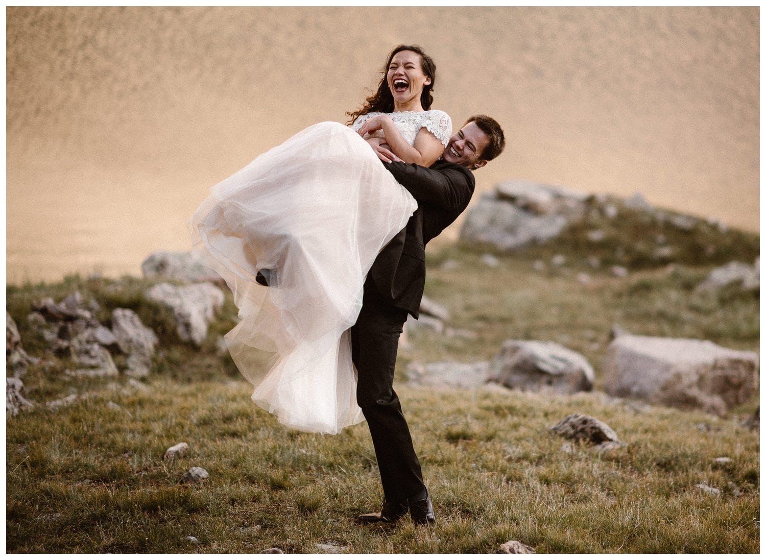 Groom lifts up bride in a grassy meadow and they are both laughing. 