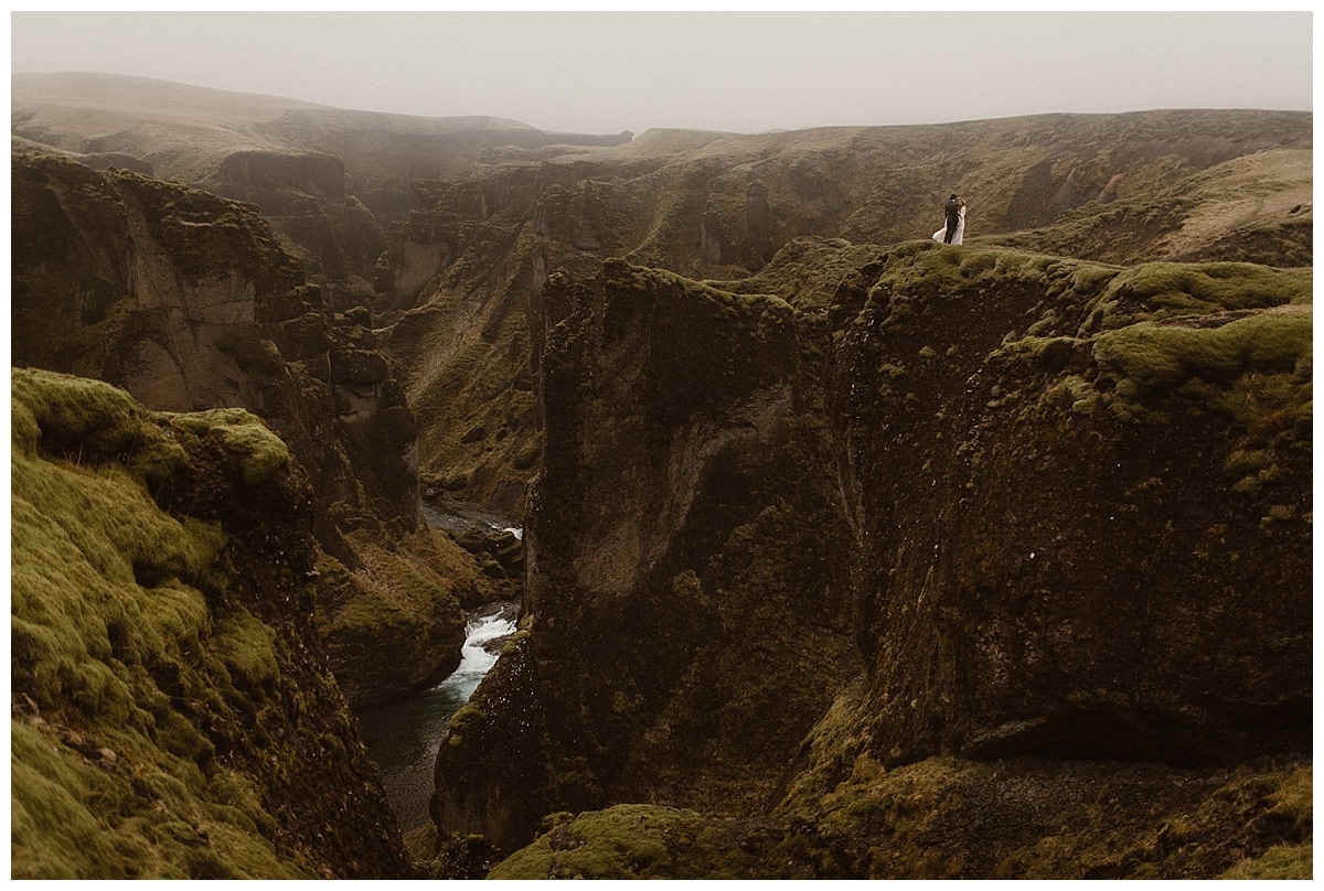 bride and groom standing on mossy green cliff in Iceland, they are far off in the distance.