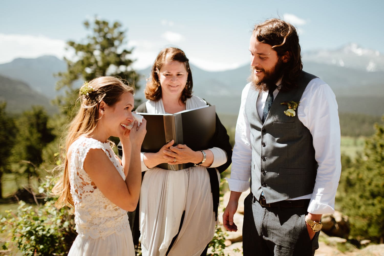 Bride wipes away tear while groom smiles at her during intimate elopement ceremony at 3M Curve in Rocky Mountain National Park, Colorado. 