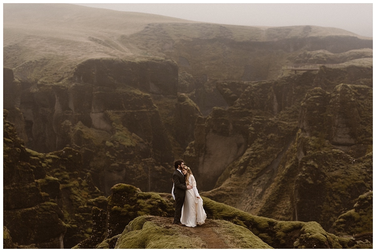 Bride and groom standing on mossy green cliff in Iceland. 