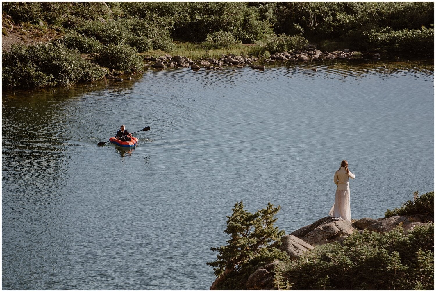 Groom in raft paddles towards bride, who is standing on island in alpine lake. 