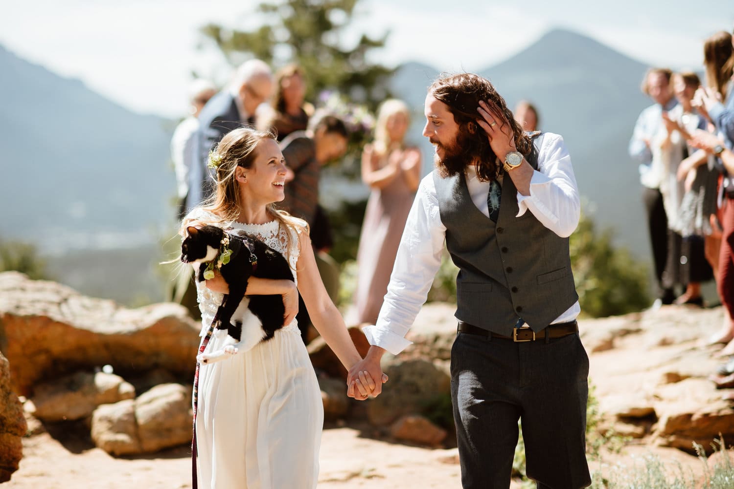 Bride and groom hold hands and smile at each other on their elopement day at 3M Curve in Rocky Mountain National Park, Colorado. The bride is holding a black and white cat. 