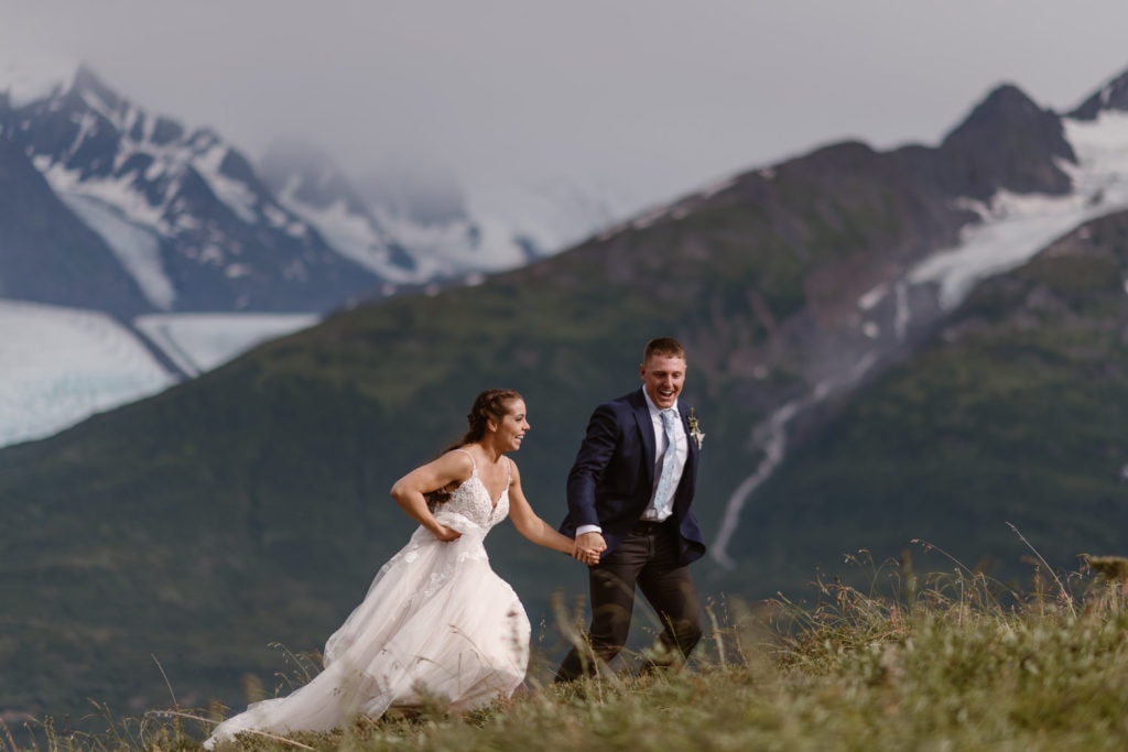 Bride and groom run through a meadow in Alaska, laughing and holding hands, with snow-capped peaks behind them 