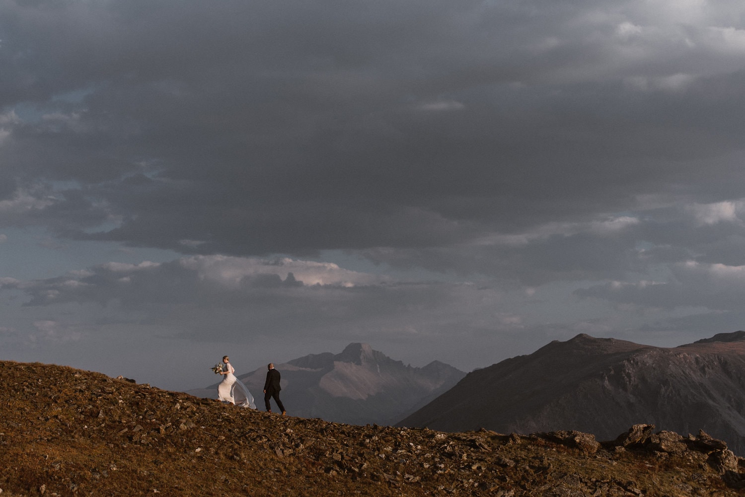 Bride and groom walking along a mountain on their elopement day. The sky is cloudy and there are mountains in the background. 