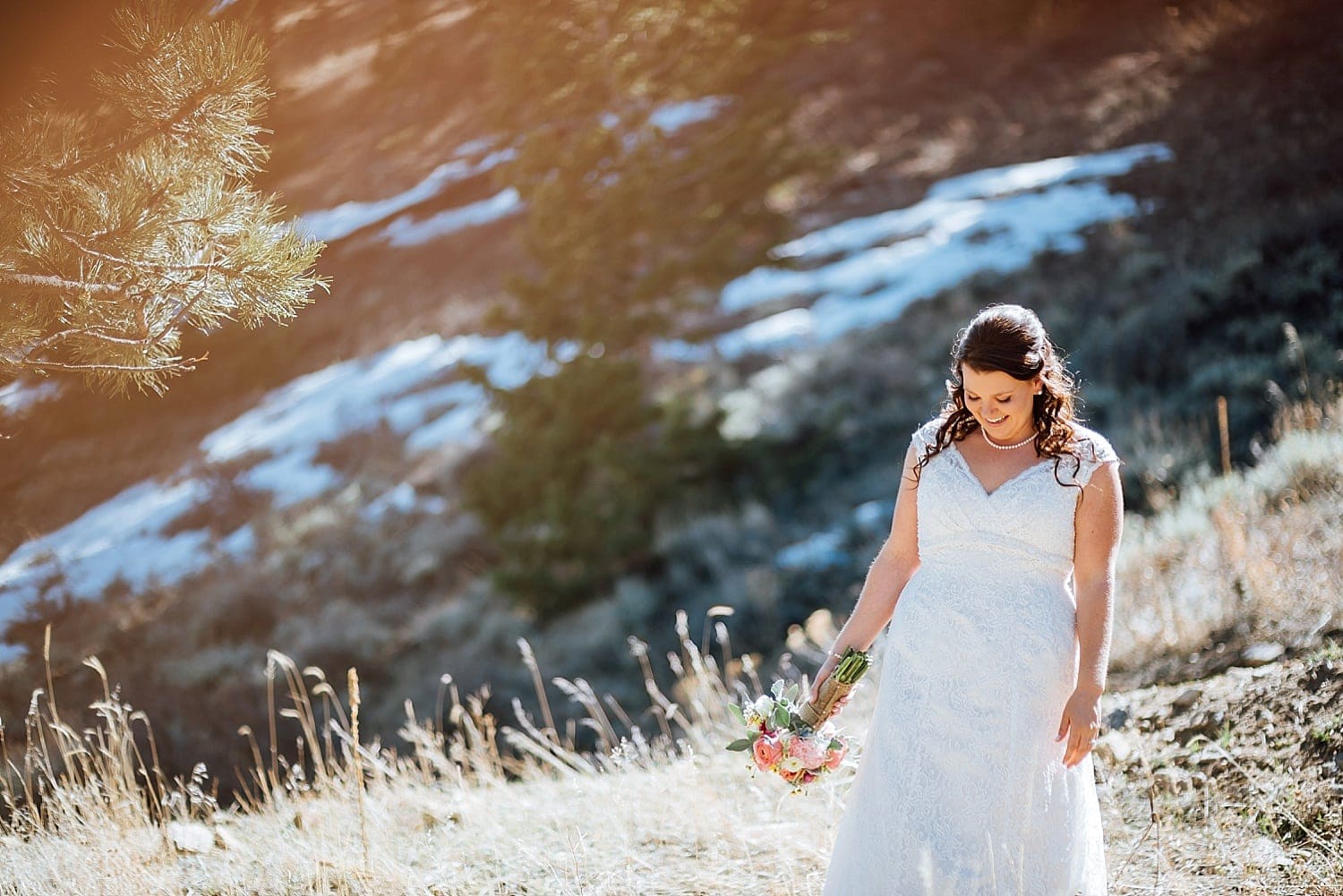 Bride walks through meadow on her elopement day in Estes Park, Colorado. She is wearing a white lace dress, a pearl necklace, with her hair curled, and holding a bouquet. 