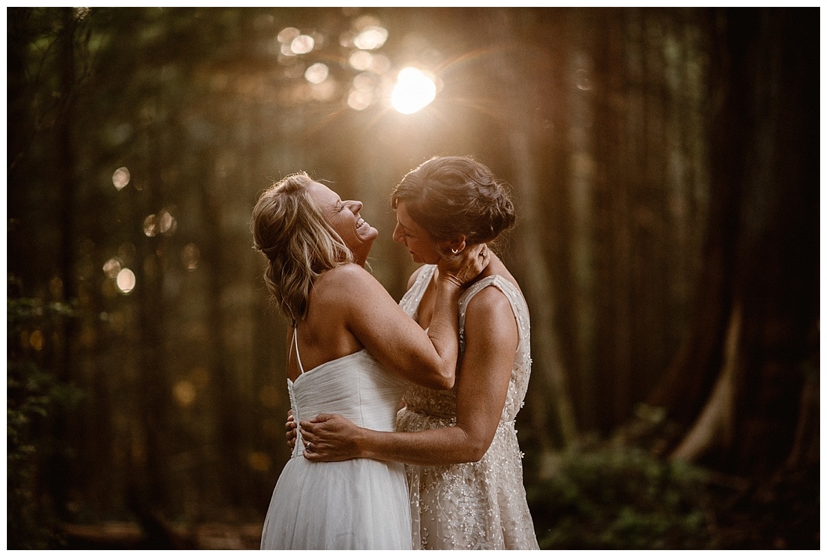Two brides embrace and laugh in a forest on their elopement day in Tofino, Canada. 