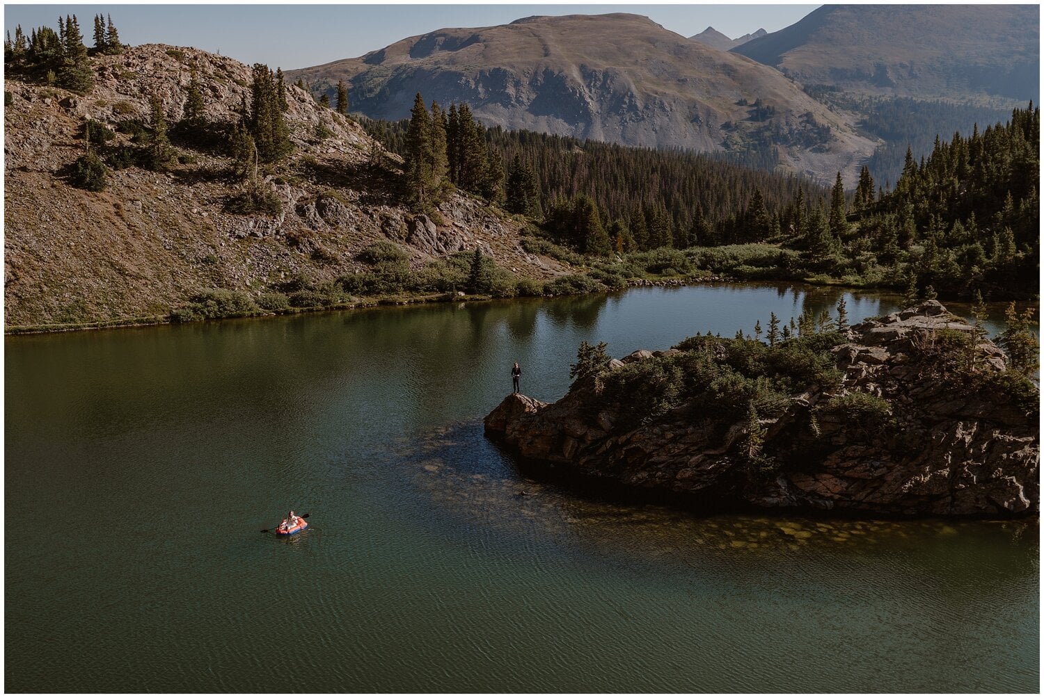 Bride paddles in raft as groom stands on island in alpine lake in Buena Vista, Colorado. 