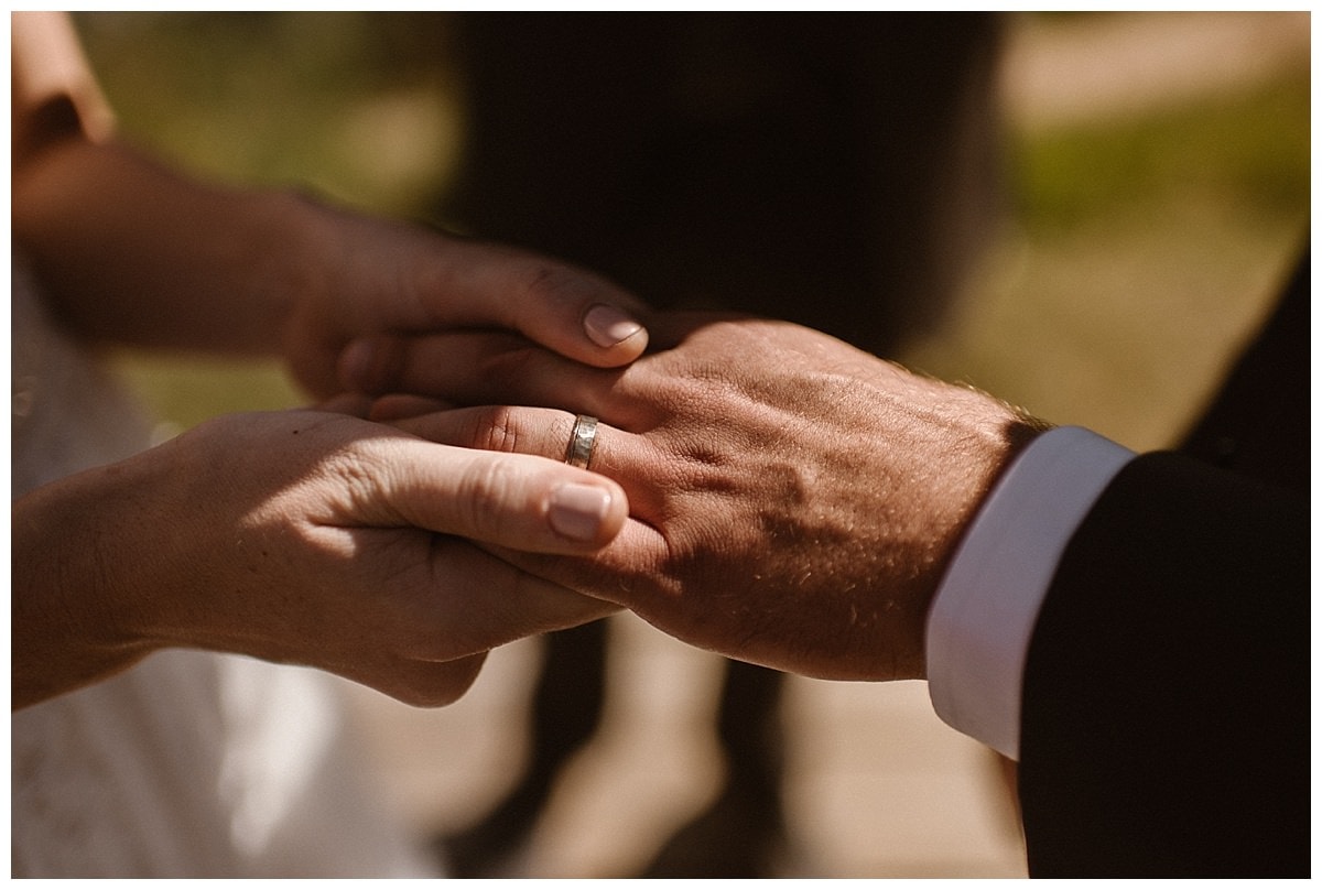 Close-up of bride holding grooms hand. Light is shining on on the ring on his ring finger. 