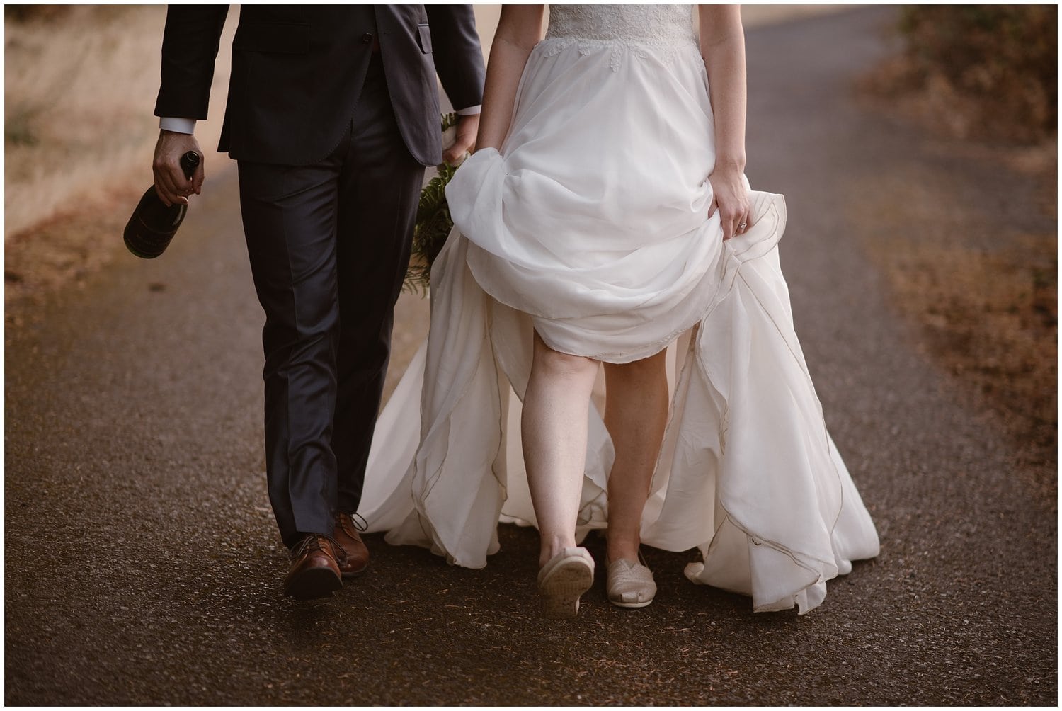 Bride and groom walking along a road together. Bride it holding up the skirt of her dress to show her shoes. 