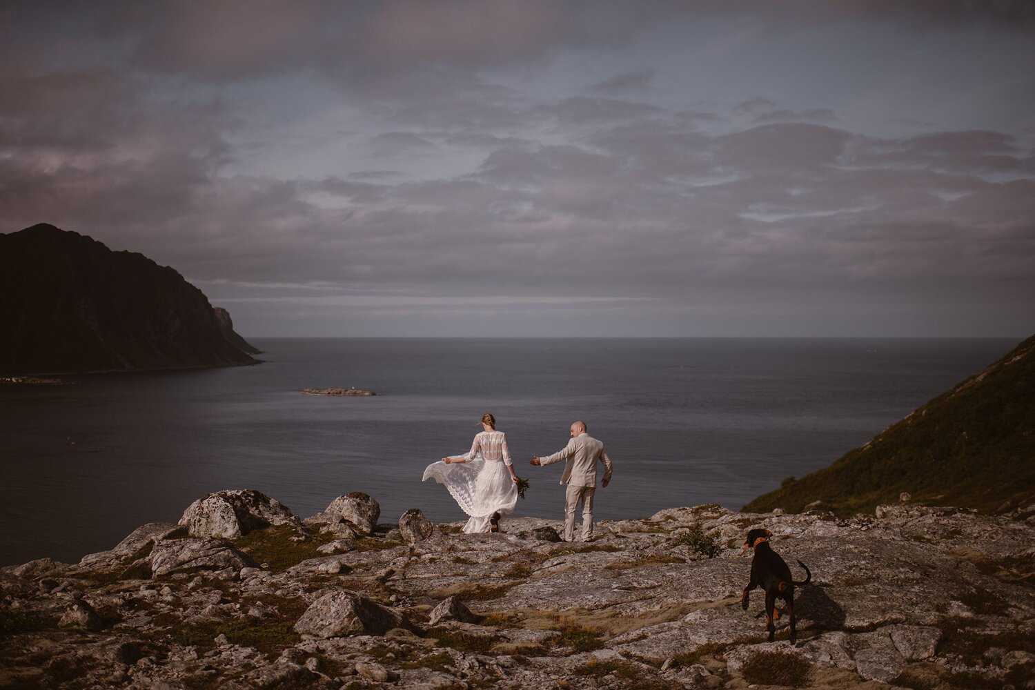 Bride and groom walk along coastal rocks with their dog, on their elopement day in Norway. 