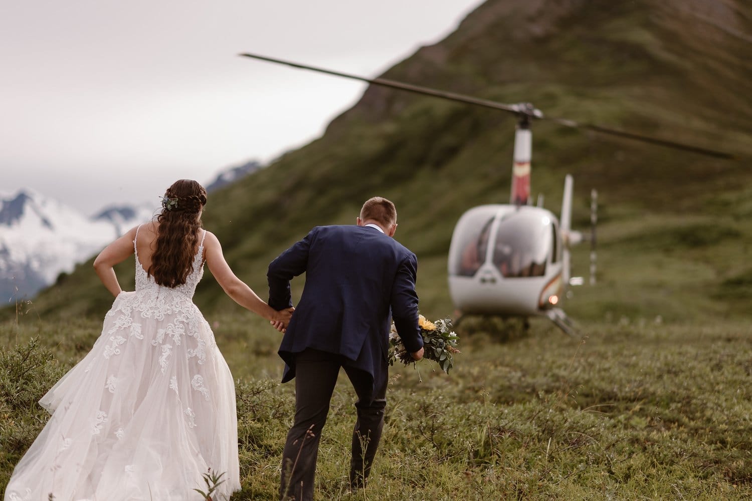 Bride and groom hold hands and walk towards helicopter in Alaska. 