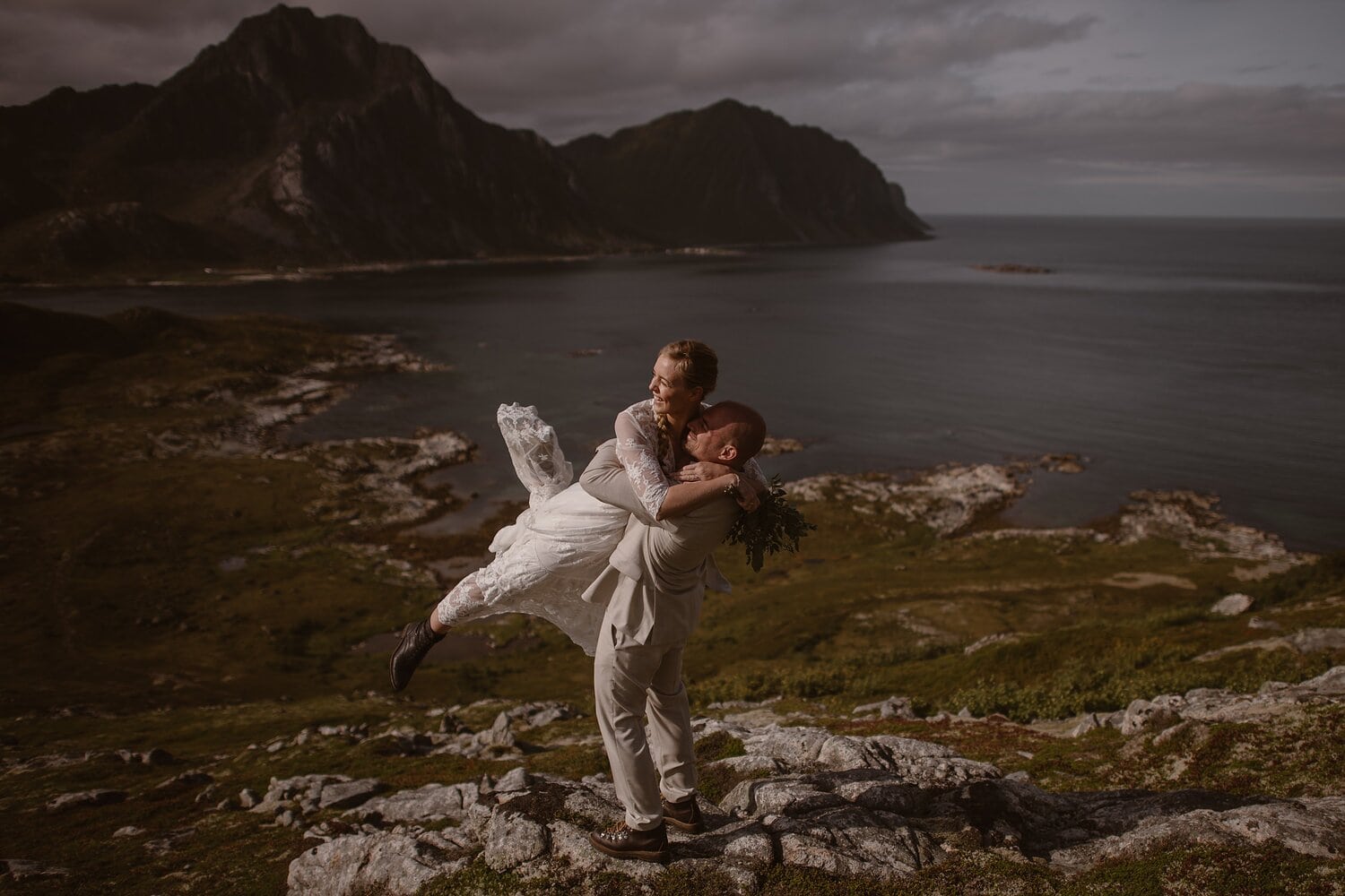 Groom lifts up bride and they are both laughing. There is a beach in the background in Norway.