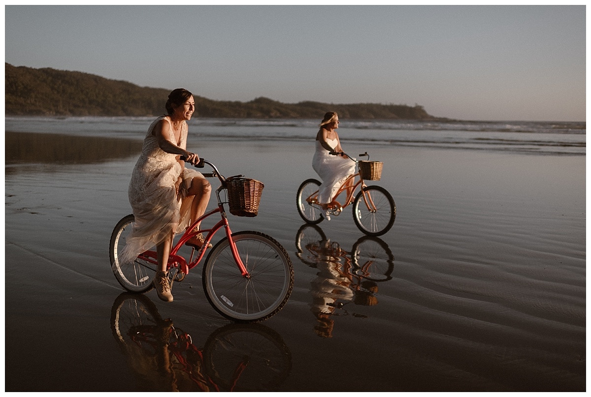 Two brides ride cruiser bikes on the beach in Tofino, Canada. 