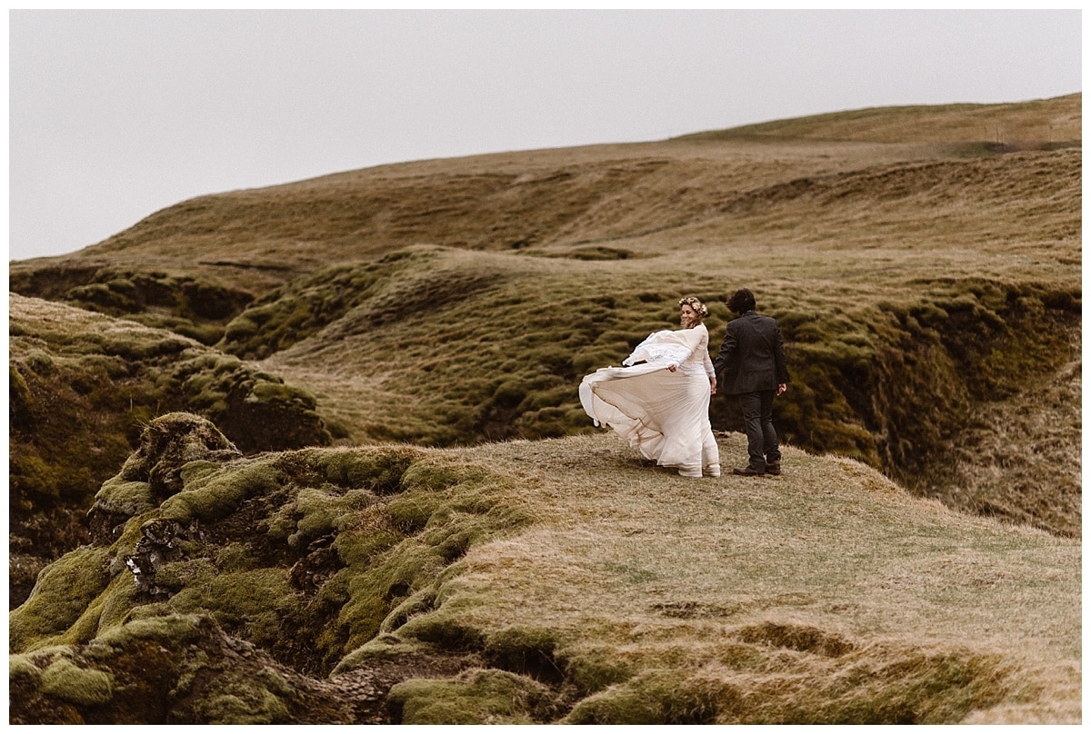 Bride and groom stand on the edge of a cliff in Iceland. The bride's white dress is flowing behind her. 