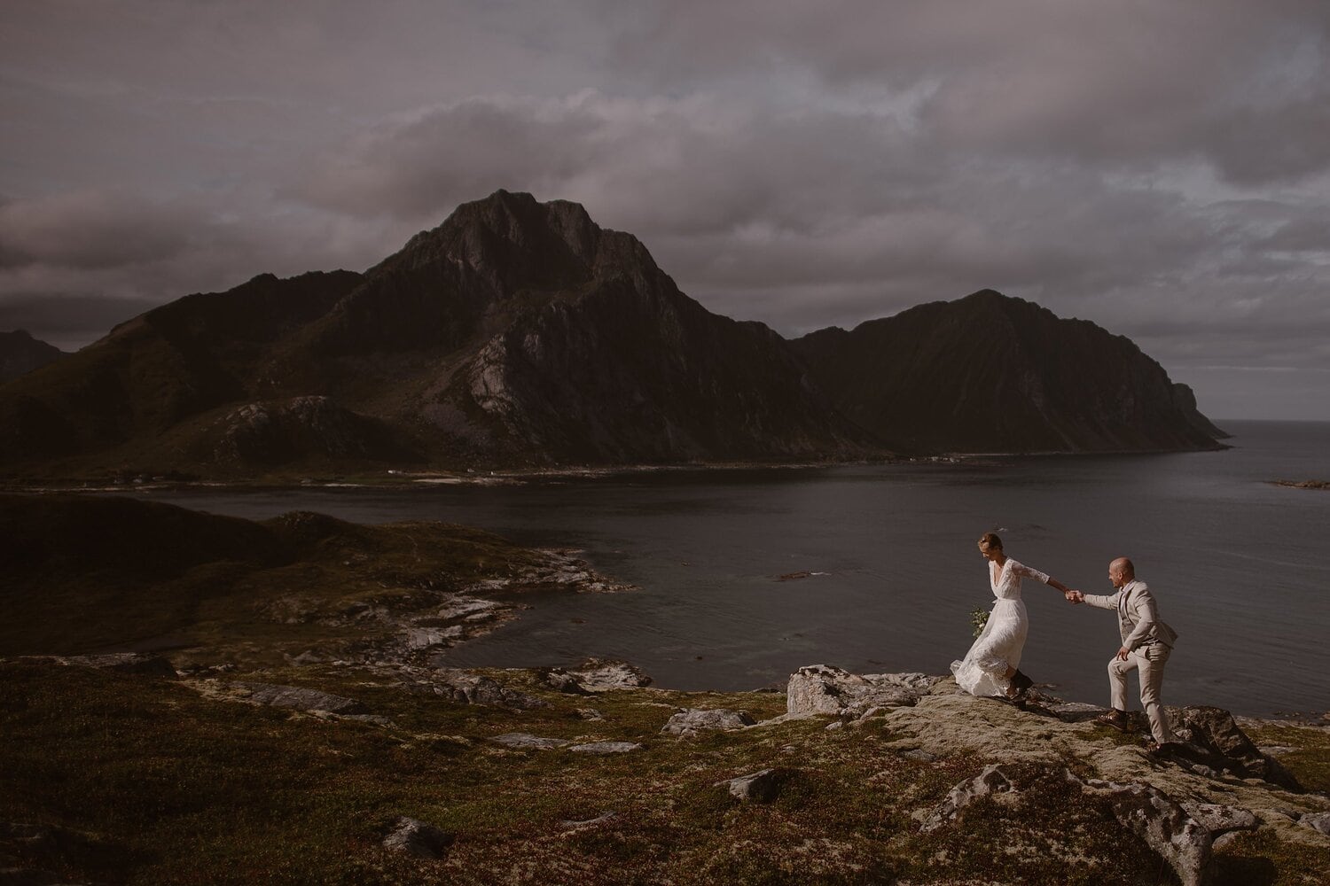 Bride and groom hold hands and hike along coast rocks at the Lofoten Islands in Norway.