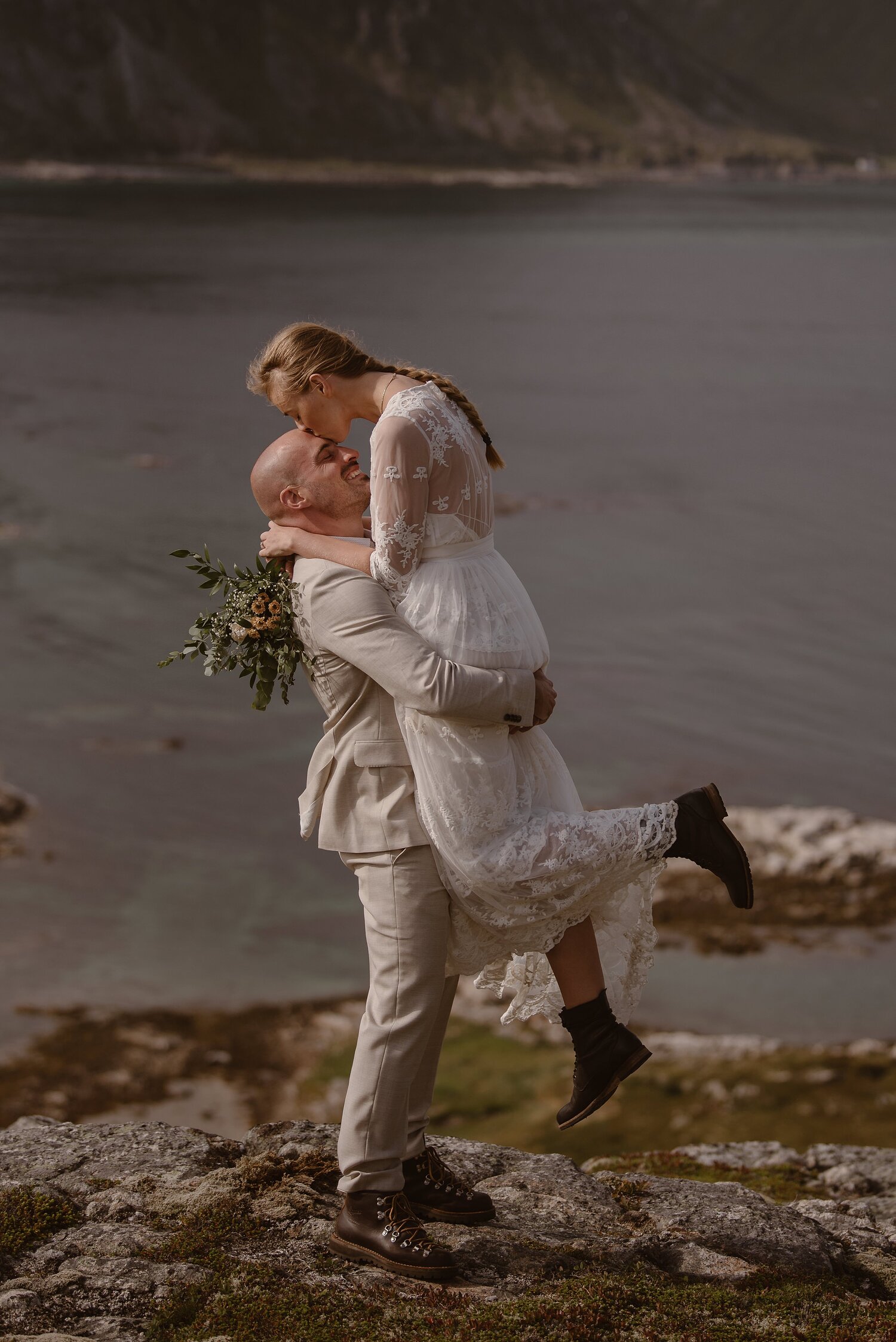 Groom lifts up bride and she kisses him on the forehead. There is a beach in the background and it's their elopement day at the Lofoten Islands in Norway.