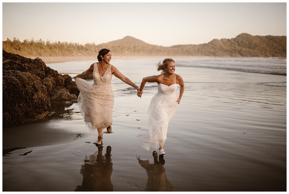 Two brides hold hands and run across the beach in Tofino, Canada. 