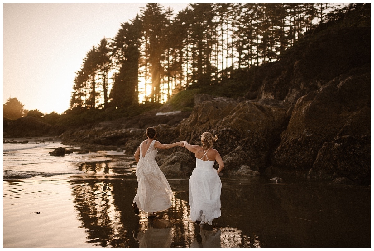 Two brides hold hands and run across the beach on their elopement day  in Tofino, Canada. 