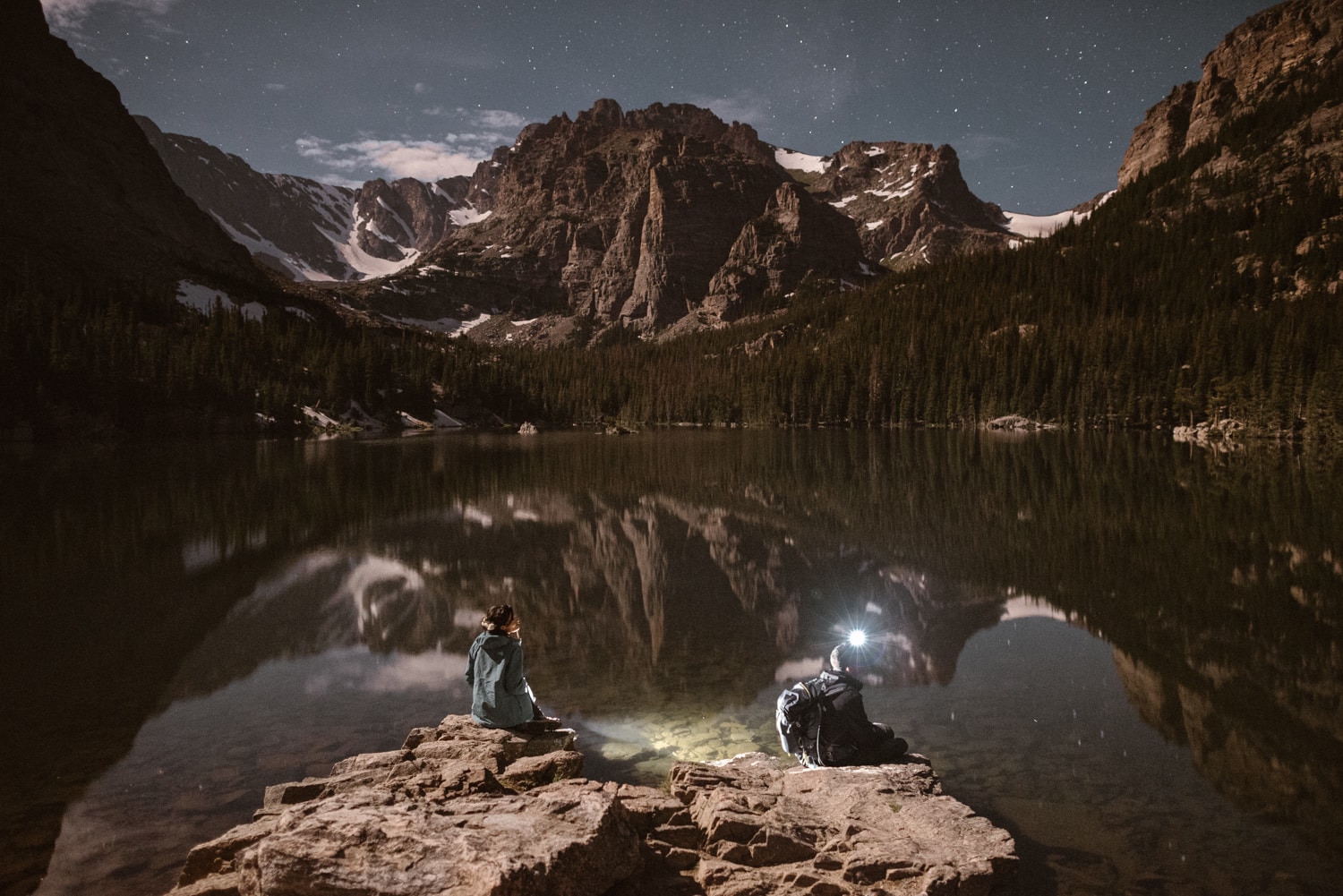 bride and groom sitting on rocks at the edge of an alpine lake at night. There are stars in the sky, and snow-covered mountains in the background. 