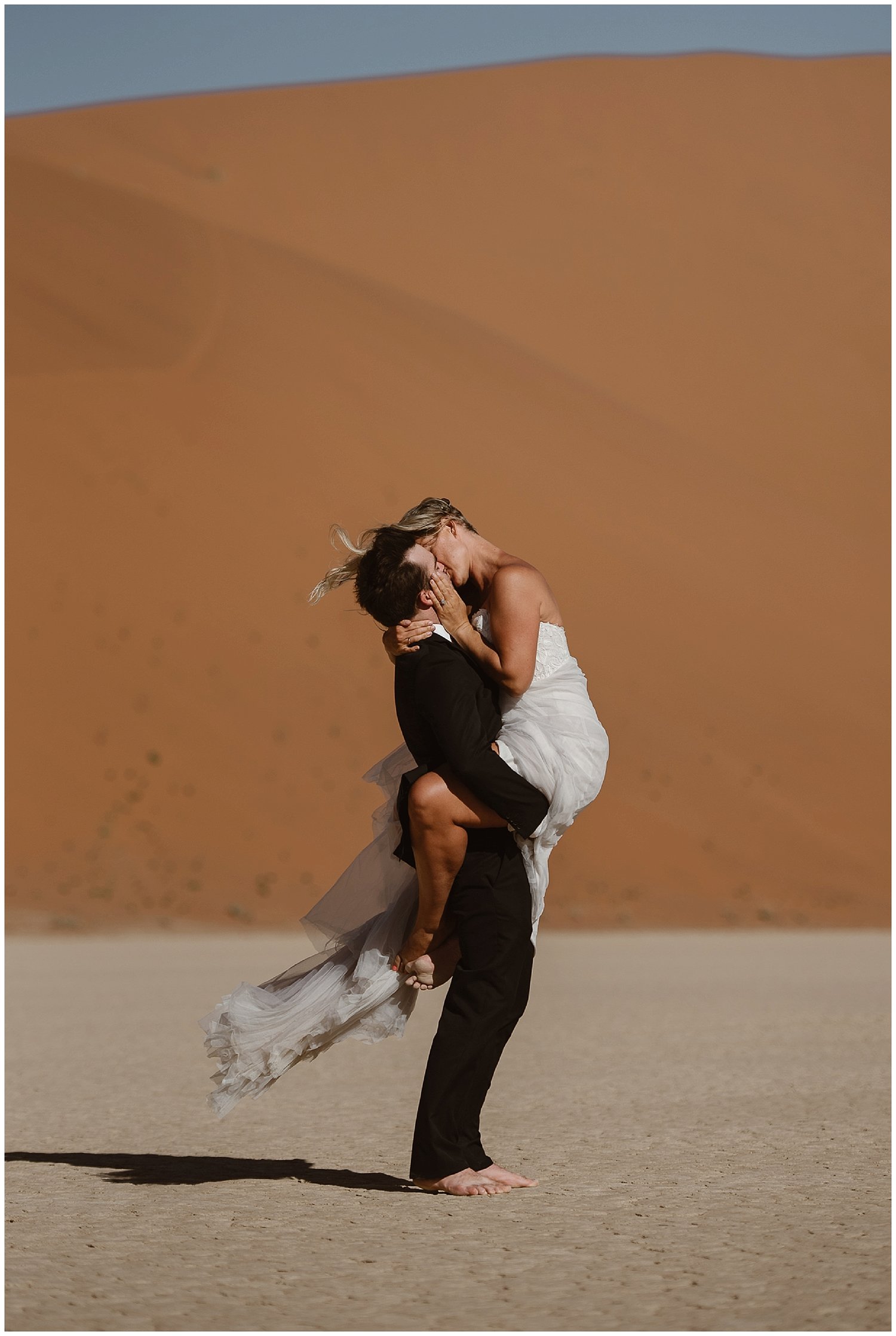 Groom holds bride as they kiss in desert. 