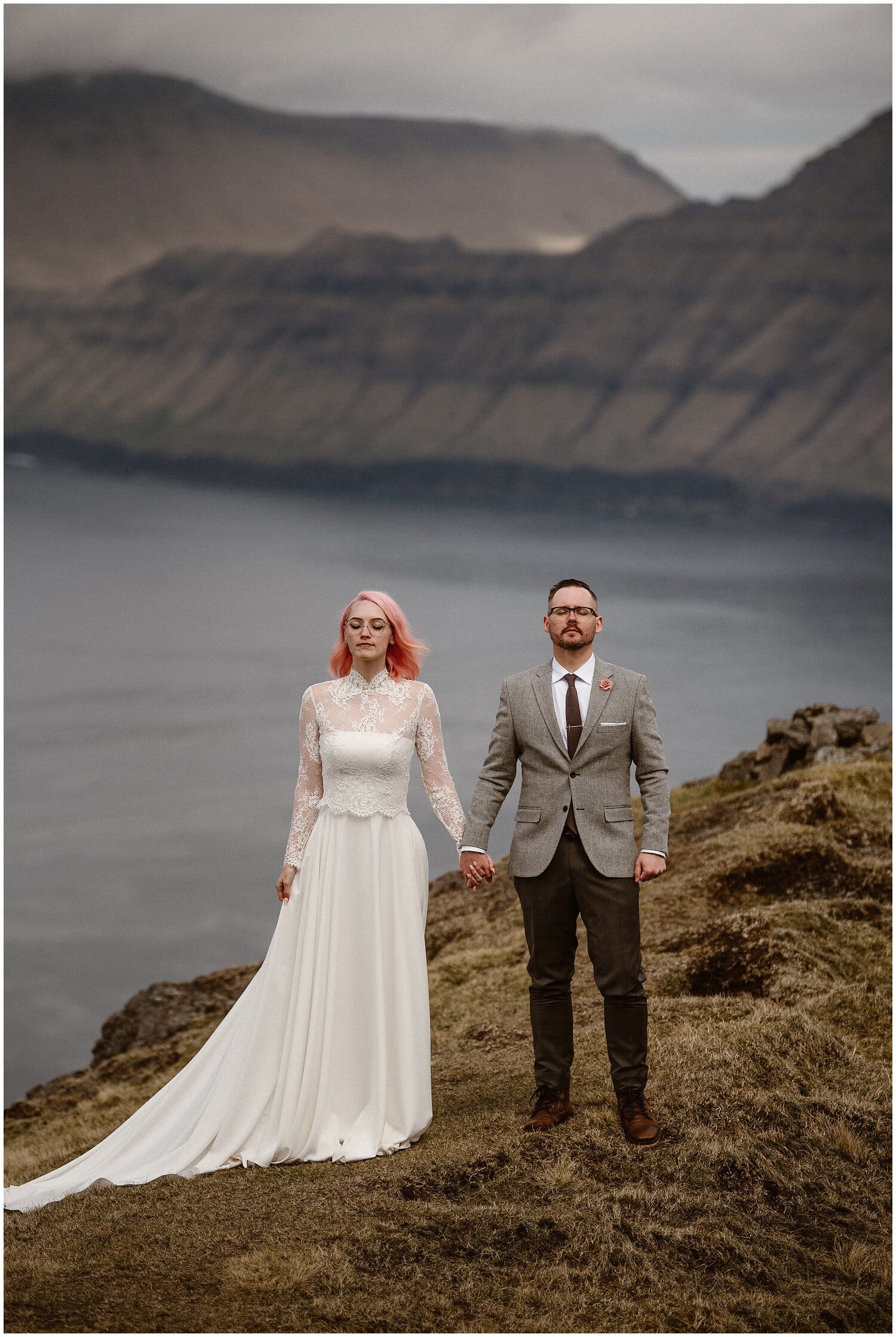 Bride and groom stand on cliff overlooking fjord in Faroe Islands. 