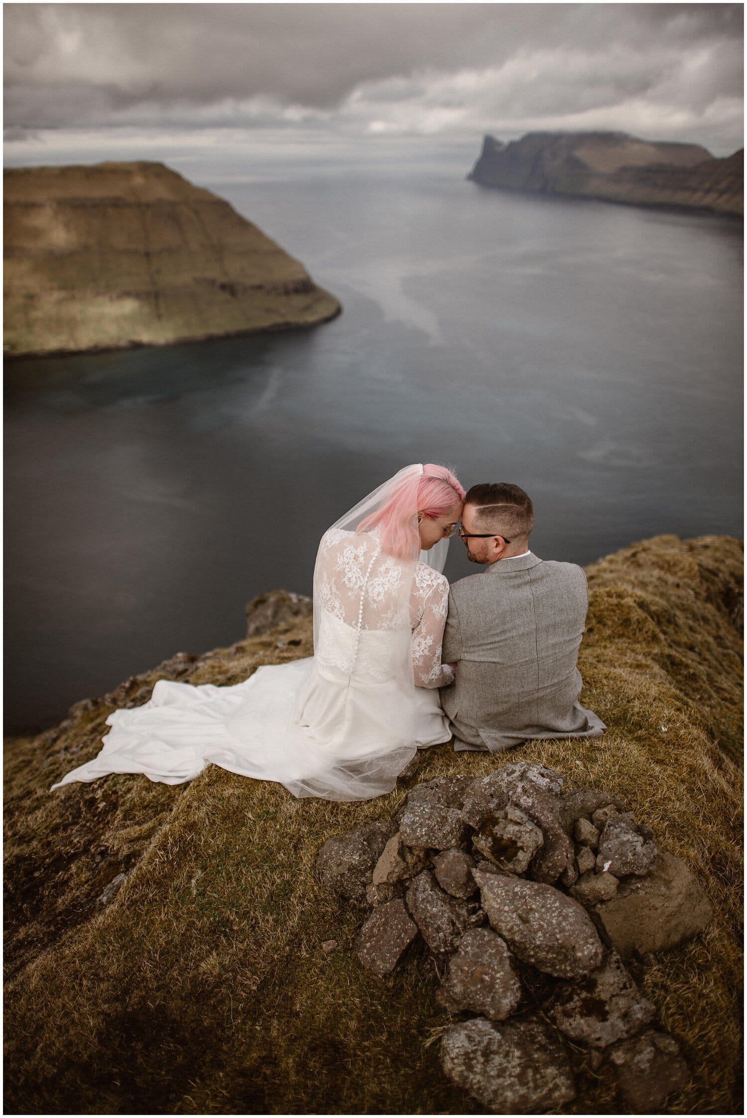 Bride and groom sit on cliff overlooking fjord in Faroe Islands. 