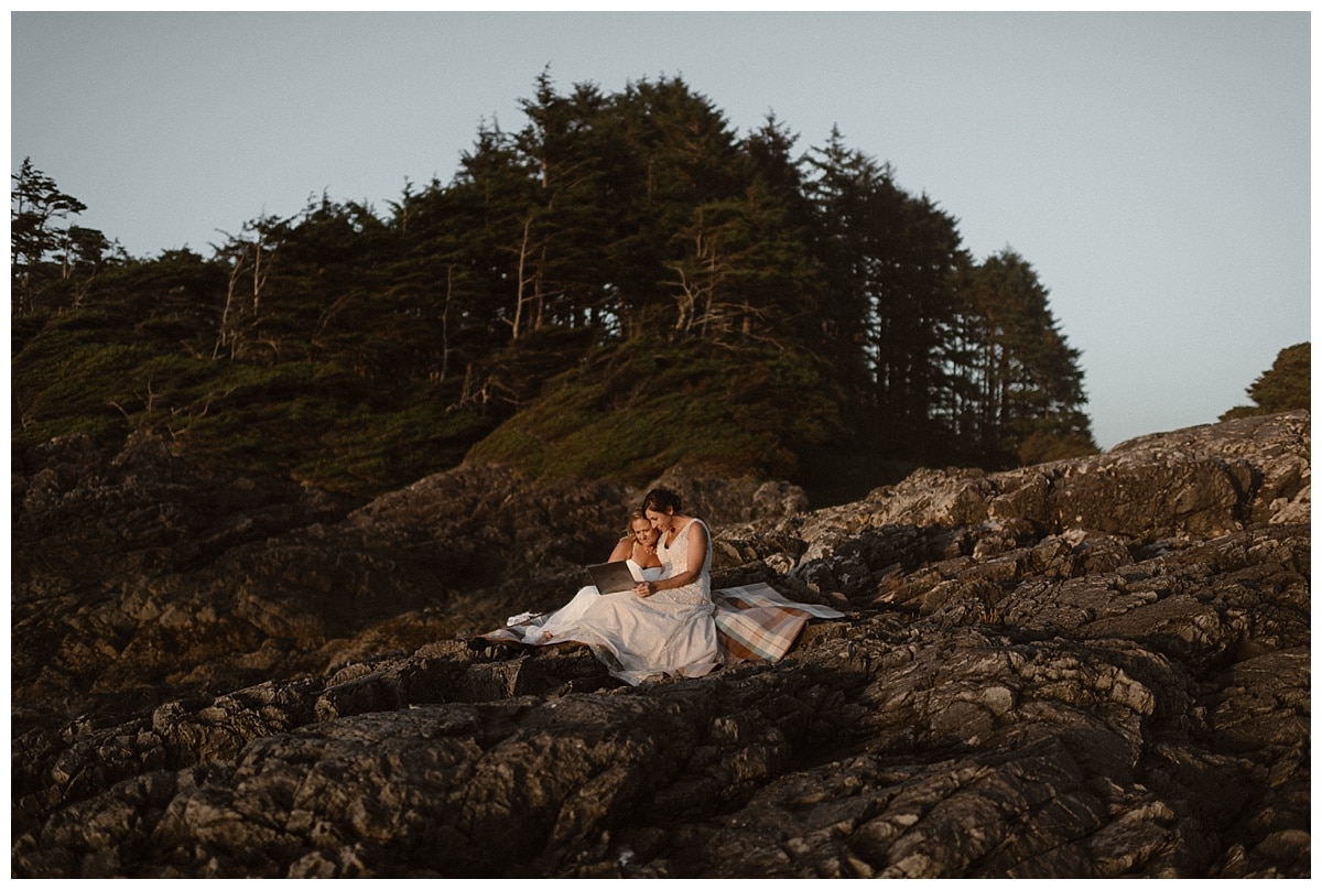 Two brides sit on a rocks near the shore and watch videos from their friends and family on their elopement day in Tofino, Canada. 