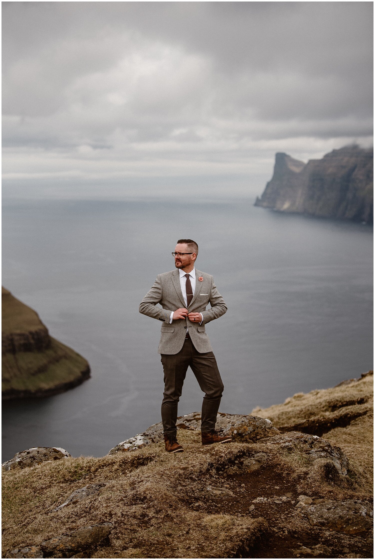 Groom stands on cliff overlooking fjord in Faroe Islands. 