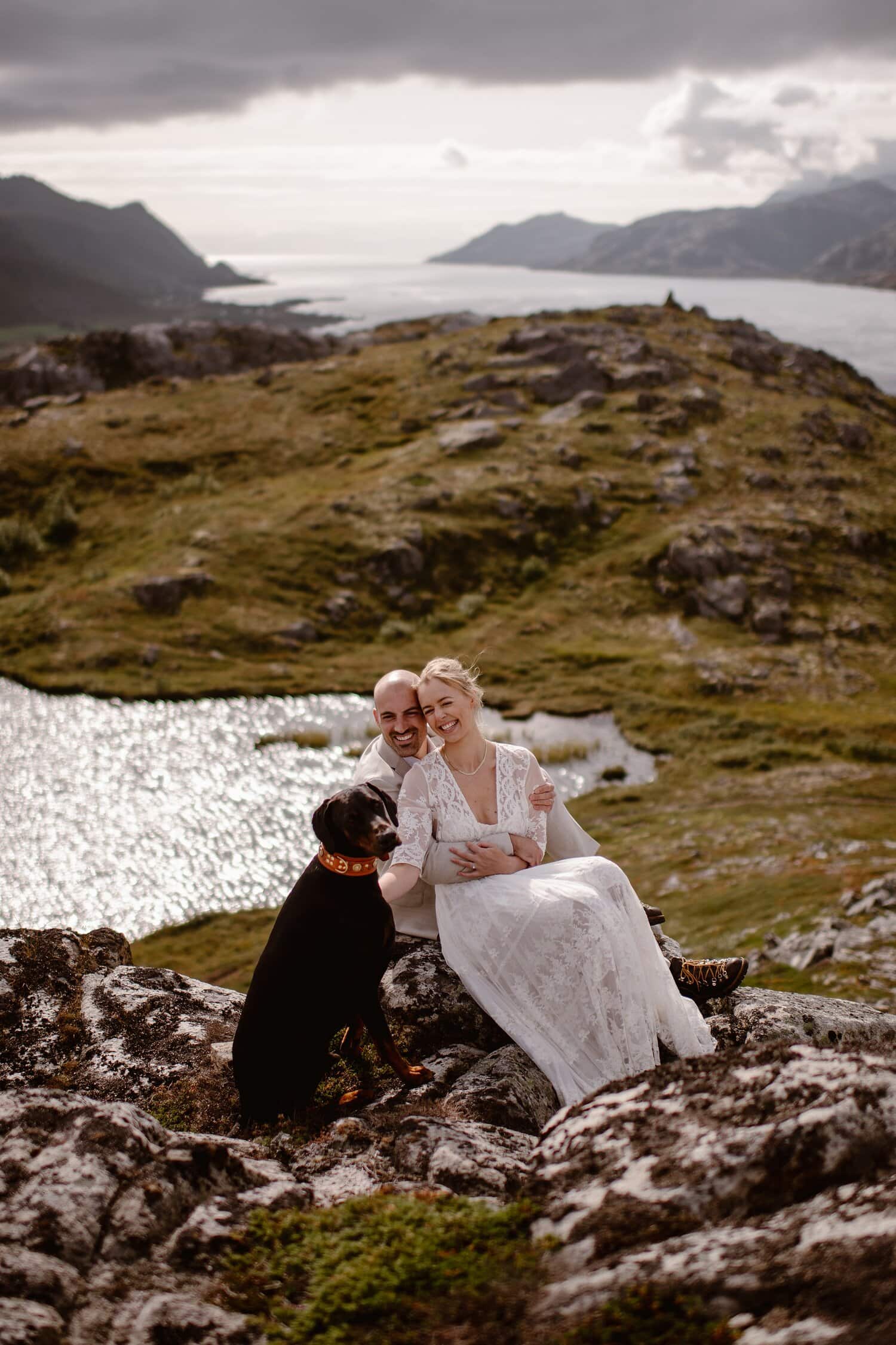 Bride and groom sit on a rock together and smile at their dog. They are at the Lofoten Islands in Norway.