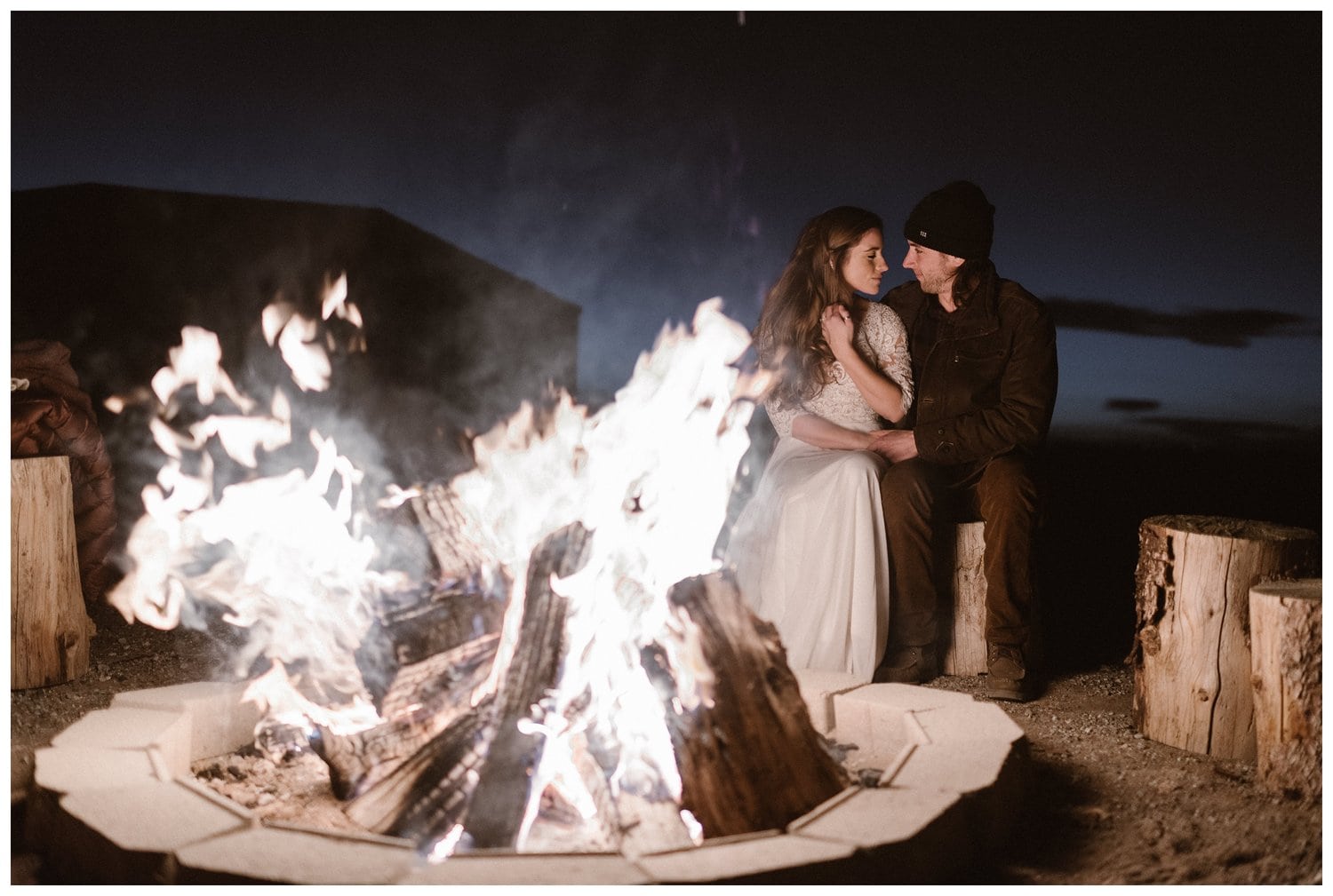 Bride and groom sit on logs next to each other with bonfire in foreground. 