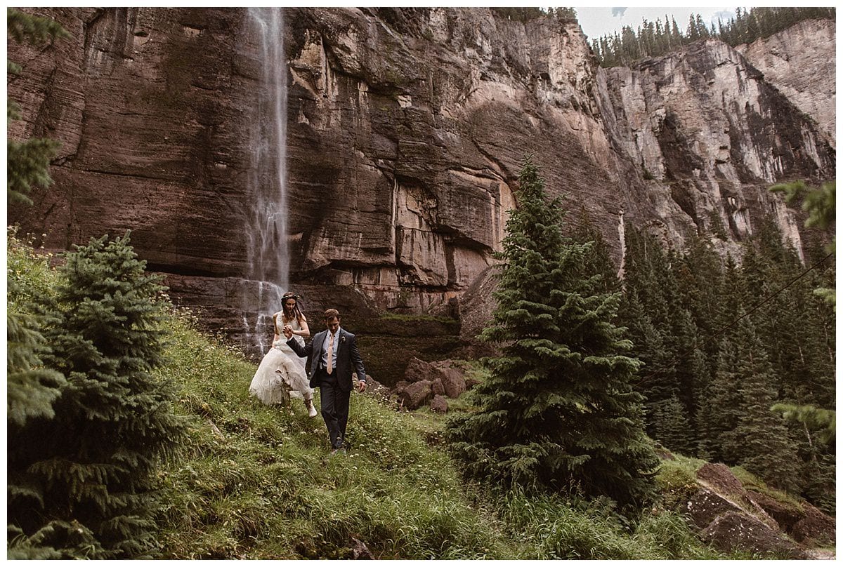 Bride and groom hold hands and walk near waterfall at Bridal Veil Falls in Telluride, Colorado. 