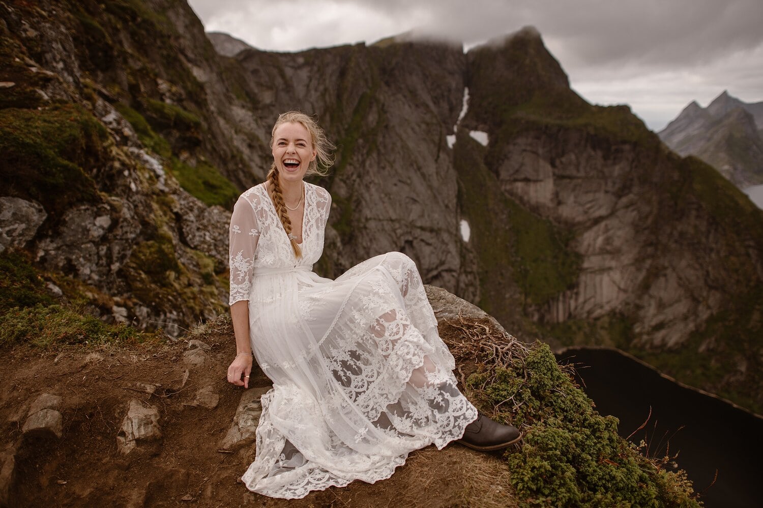 Bride sitting on a cliff and laughing. She is wearing a white, embroidered wedding dress. 