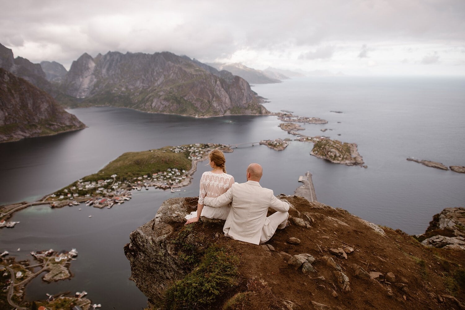 Bride and groom sit on a cliff together and look out at the water, at the Lofoten Islands in Norway.