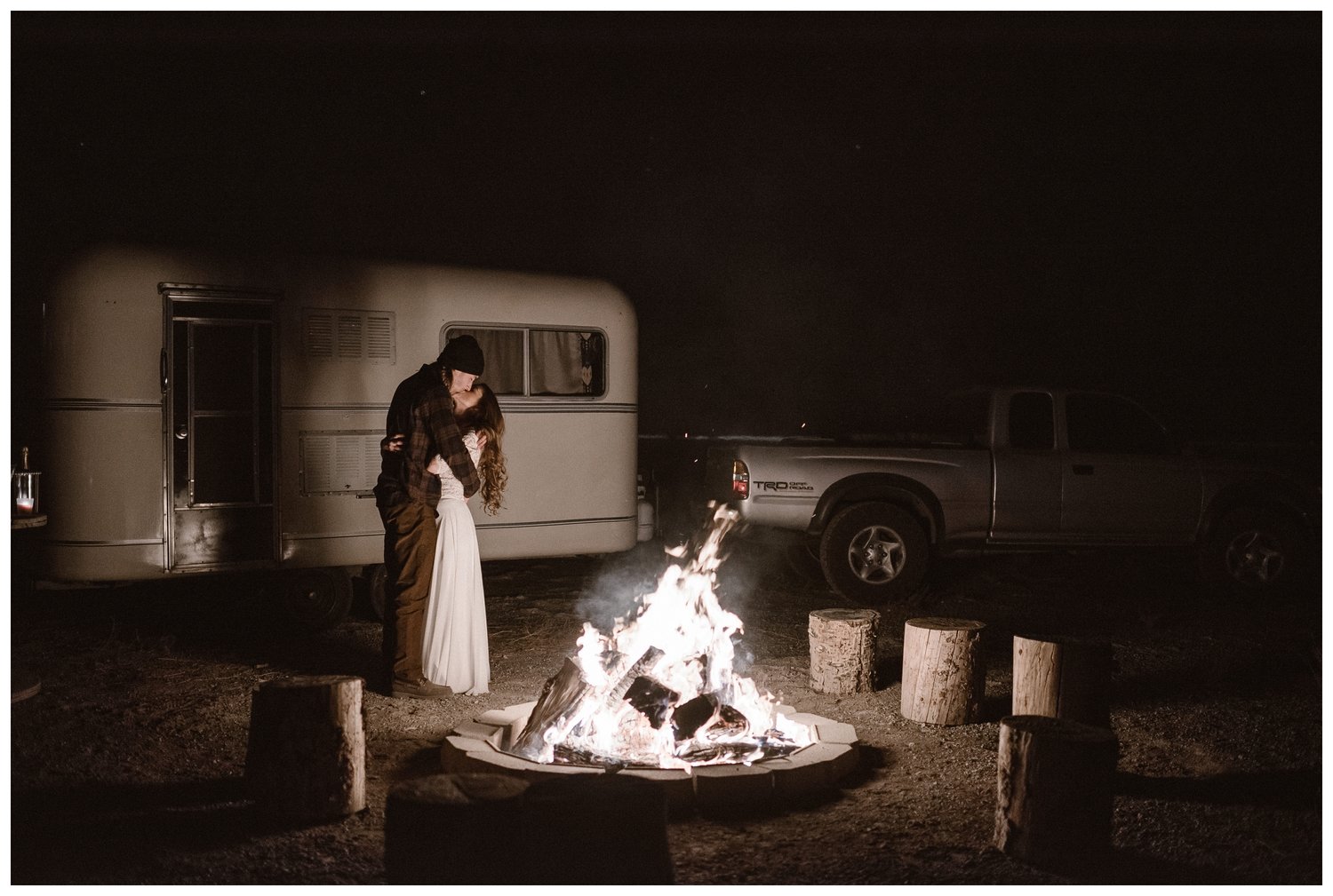 Bride and groom kiss with bonfire in foreground and camper in background. 