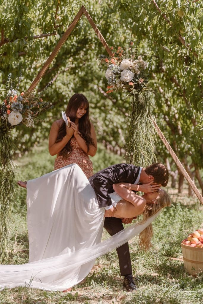 Groom dips bride for a kiss during intimate ceremony at a peach orchard in Palisade, Colorado. 