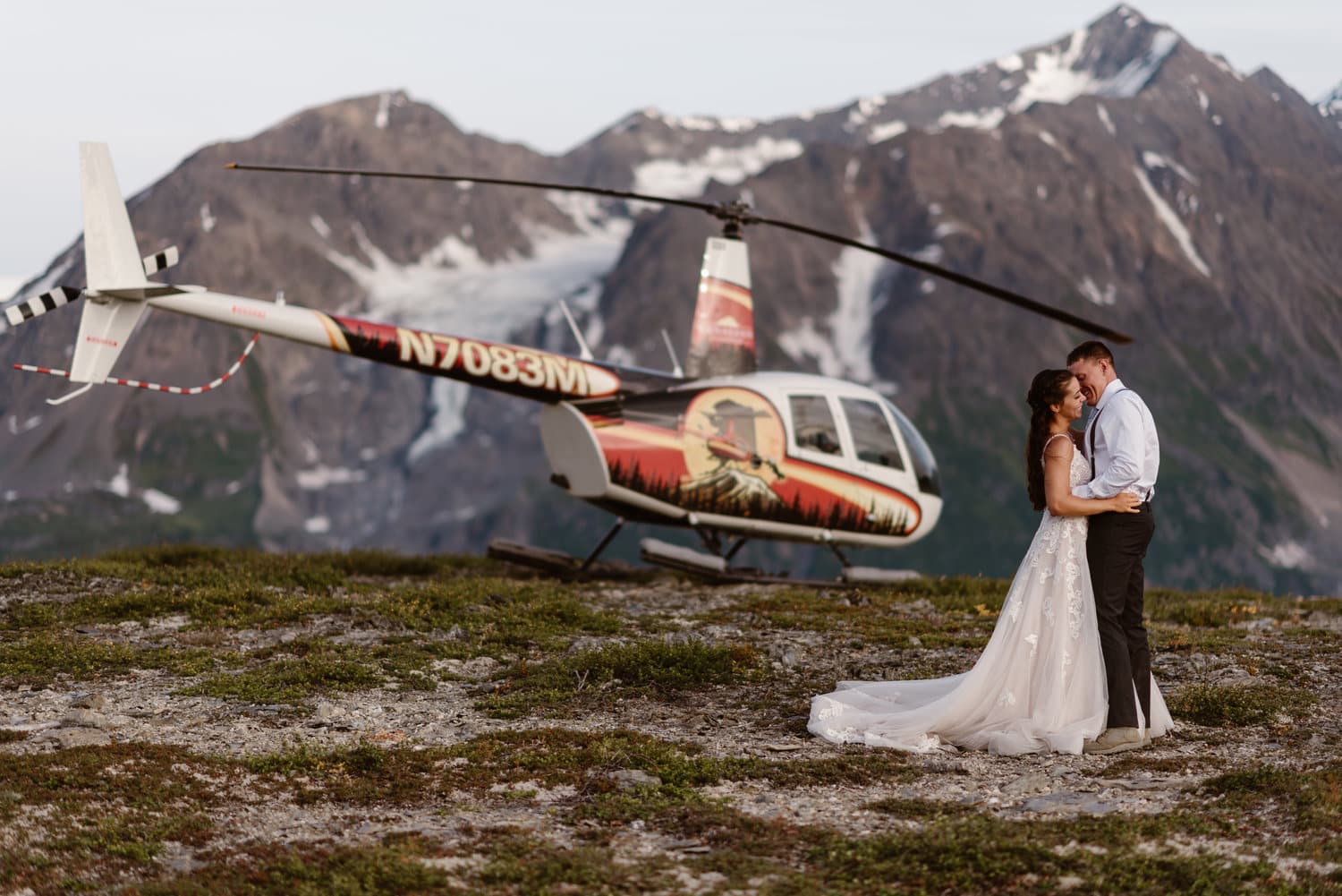 Bride and groom embrace in front of helicopter on their elopement day in Alaska. There are snow-capped mountains in the background. 