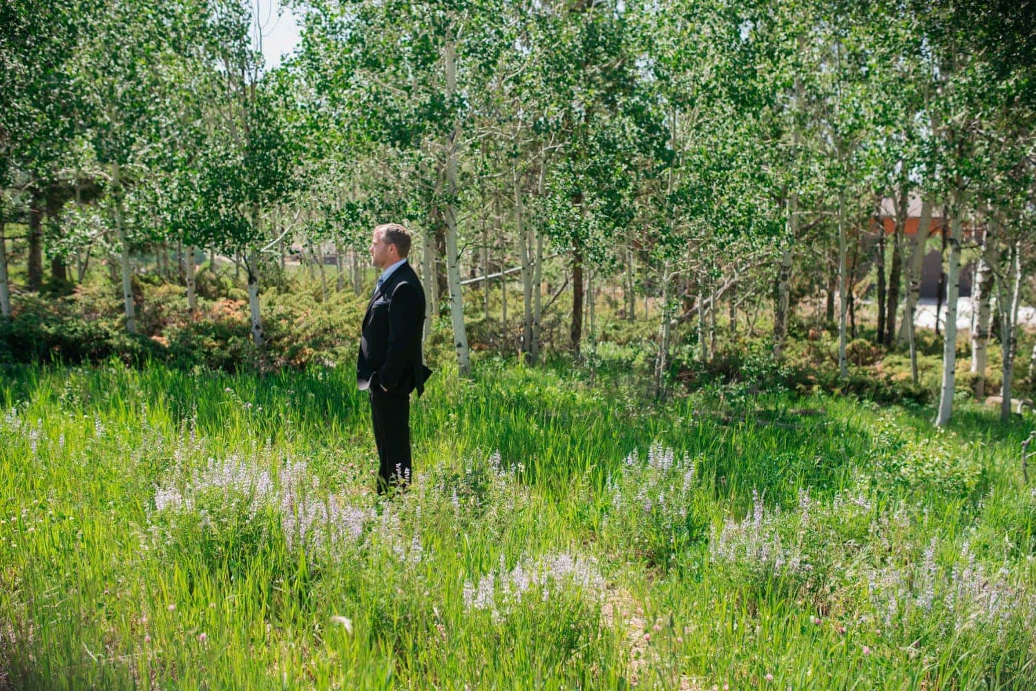 Groom standing in a meadow in Grand Lake, Colorado. There is tall green grass and wildflowers, with an aspen grove in the background. 
