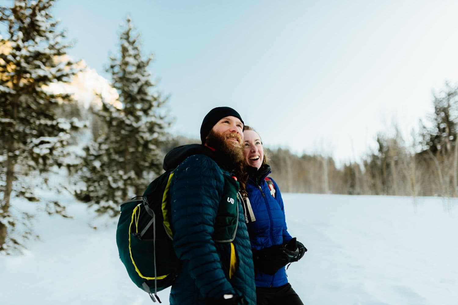 Couple standing together and smiling at Indian Peaks, in Colorado. There is snow covering the ground and forest in the background. 