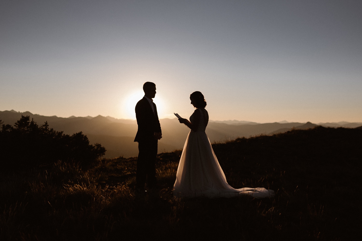 Bride reads her vows to the groom during their intimate elopement ceremony at sunrise. The sun is peeking out from behind the mountains. 