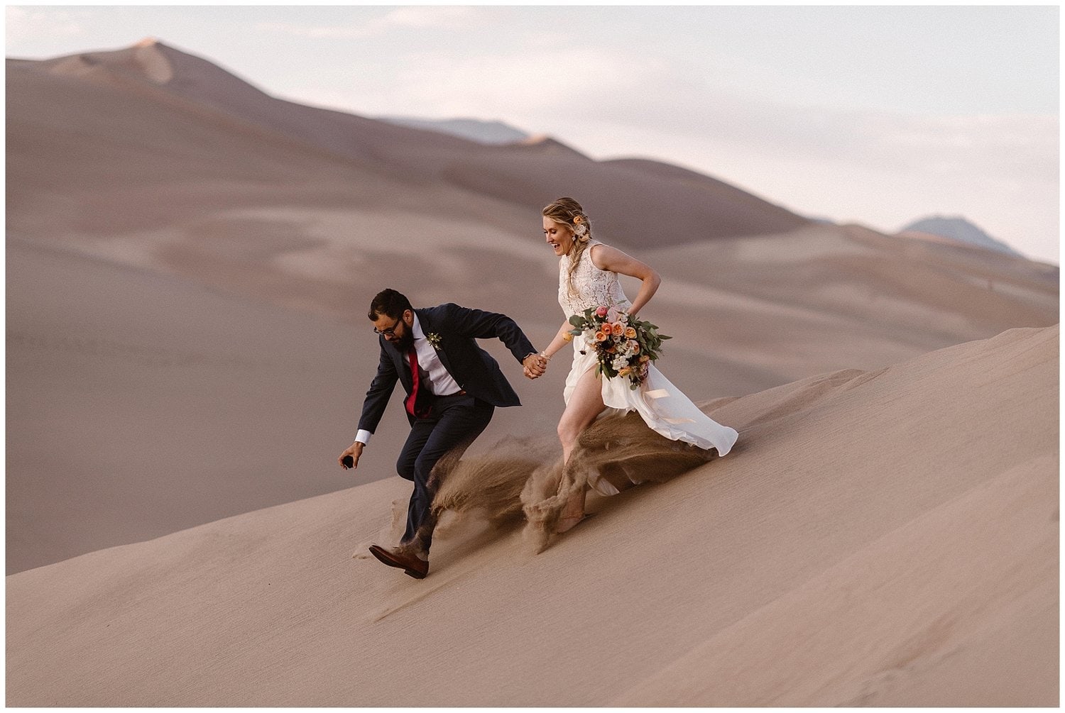 Bride and groom laughing while running down a sand dune. 