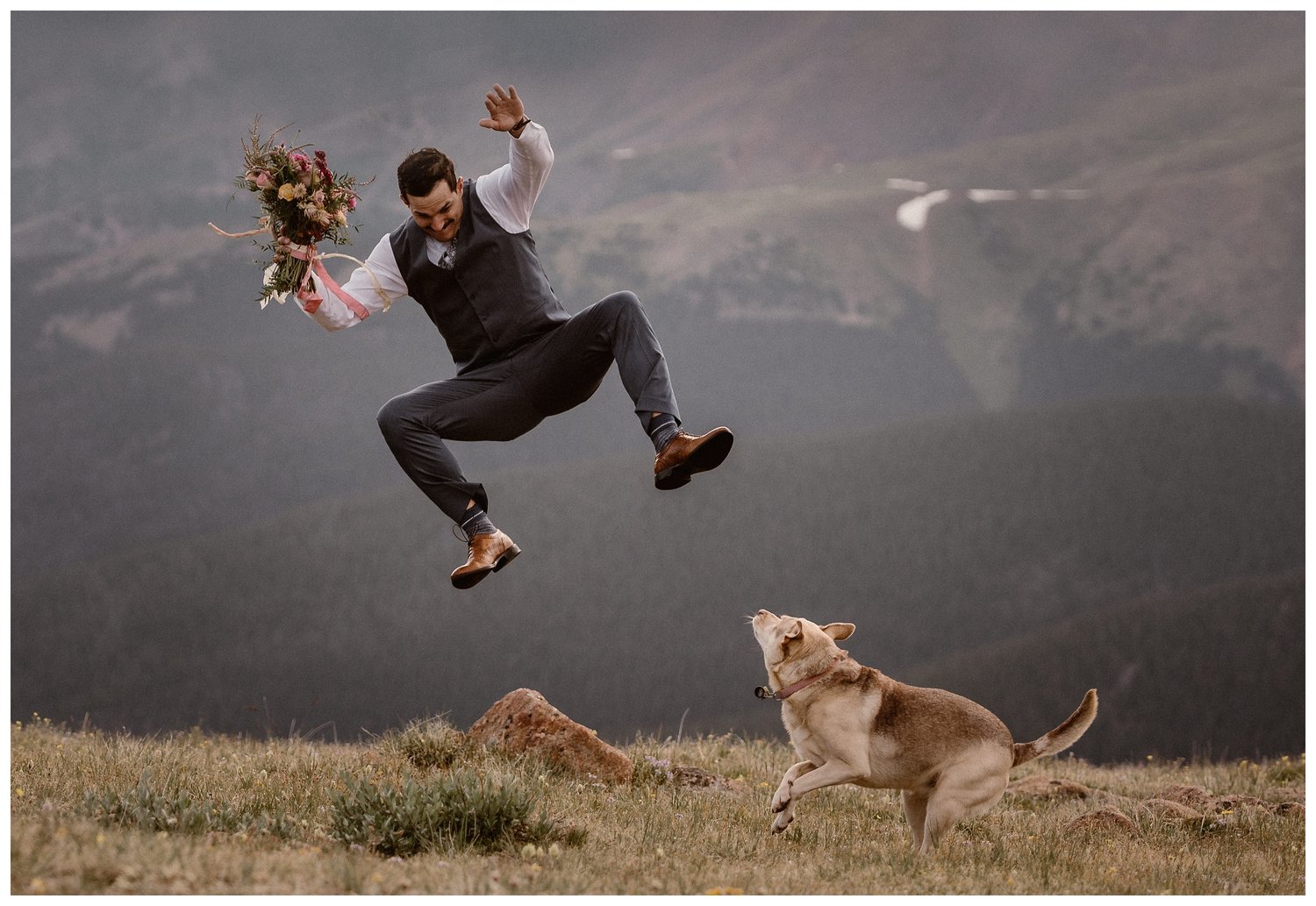 Groom jumping in the air above a dog, while holding the bride's bouquet. There are mountains in the background. 