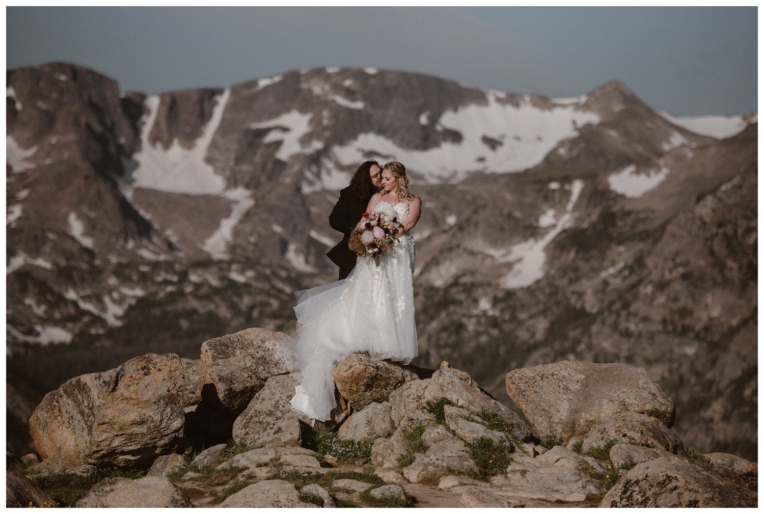 Bride and groom are standing on rocks, with mountains in the distance. Groom kisses bride on the cheek at Trail Ridge Road in Rocky Mountain National Park, Colorado. 