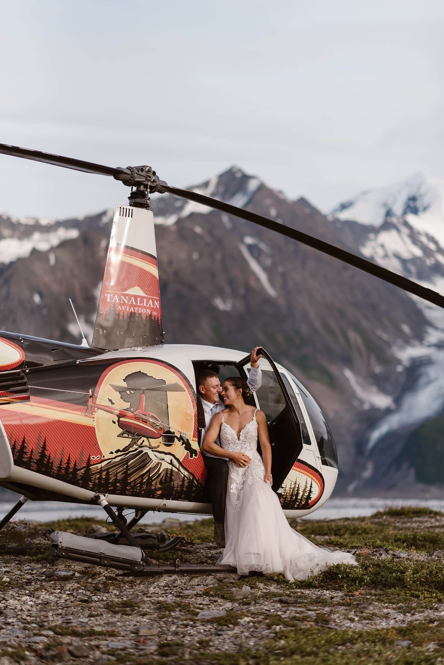Bride and groom sit in doorway of helicopter together in Alaska. There are snow-capped mountains together. 
