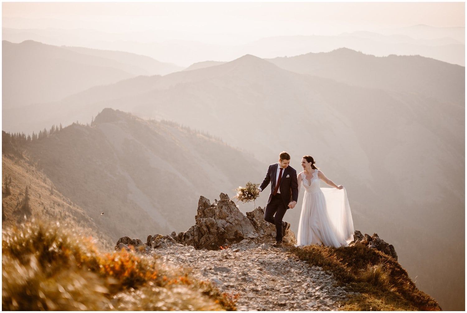 Bride and groom hold hands and walk along mountain ridge during sunrise in Washington. There are mountains in the background. 