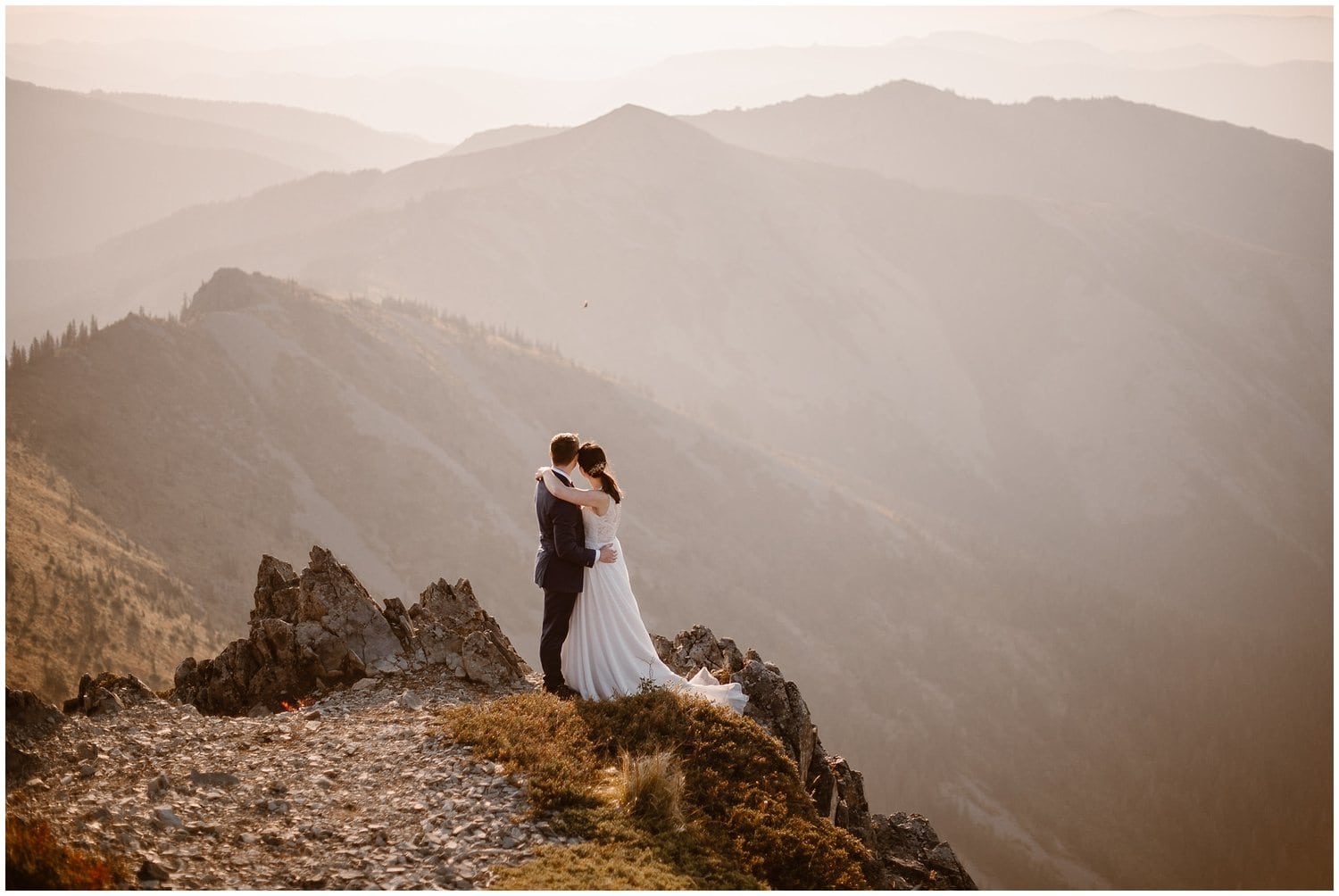 Bride and groom embrace on mountain cliff while looking out at the mountains, during sunrise in Washington. 